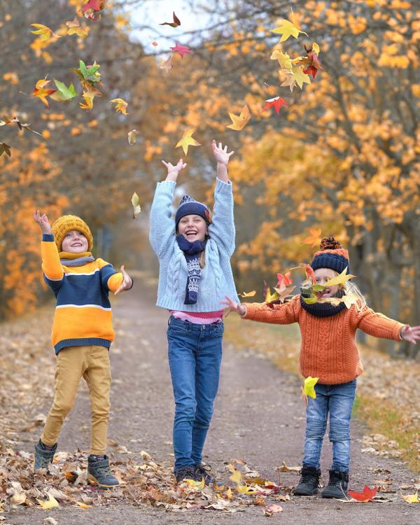 Three siblings wearing colorful hats, sweaters and scarves throw autumn leaves into the air