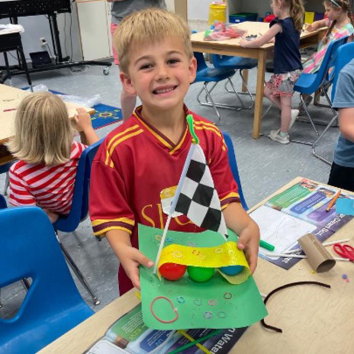 An elementary student holds up the invention prototype they made at Camp Invention with a checkered flag and multicolored spheres