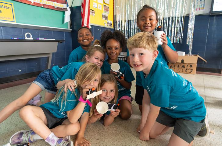 A group of Camp Invention campers smiles and holds up small robotic penguins attached to popsicle sticks and paper cups