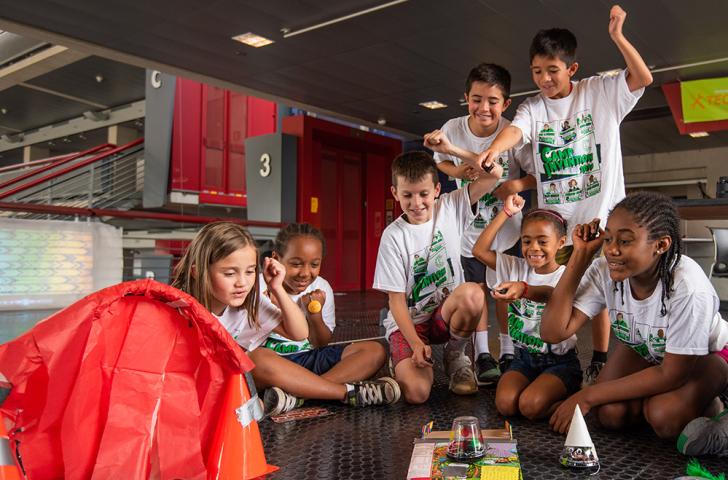 A group of Camp Invention campers cheers on a pair of cone-shaped robots moving across a classroom floor