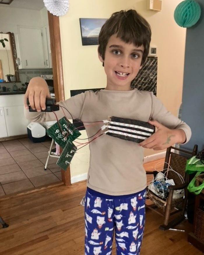 A young child smiles and holds up a light strip that they connected to a small power supply